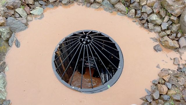A storm inlet in the road ditch — dome grate ringed with rock, winter runoff swirling down on its way to the pond.