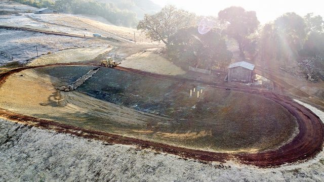 The pond just finished — the carved basin from the air in first light, its first puddle of water at the bottom.
